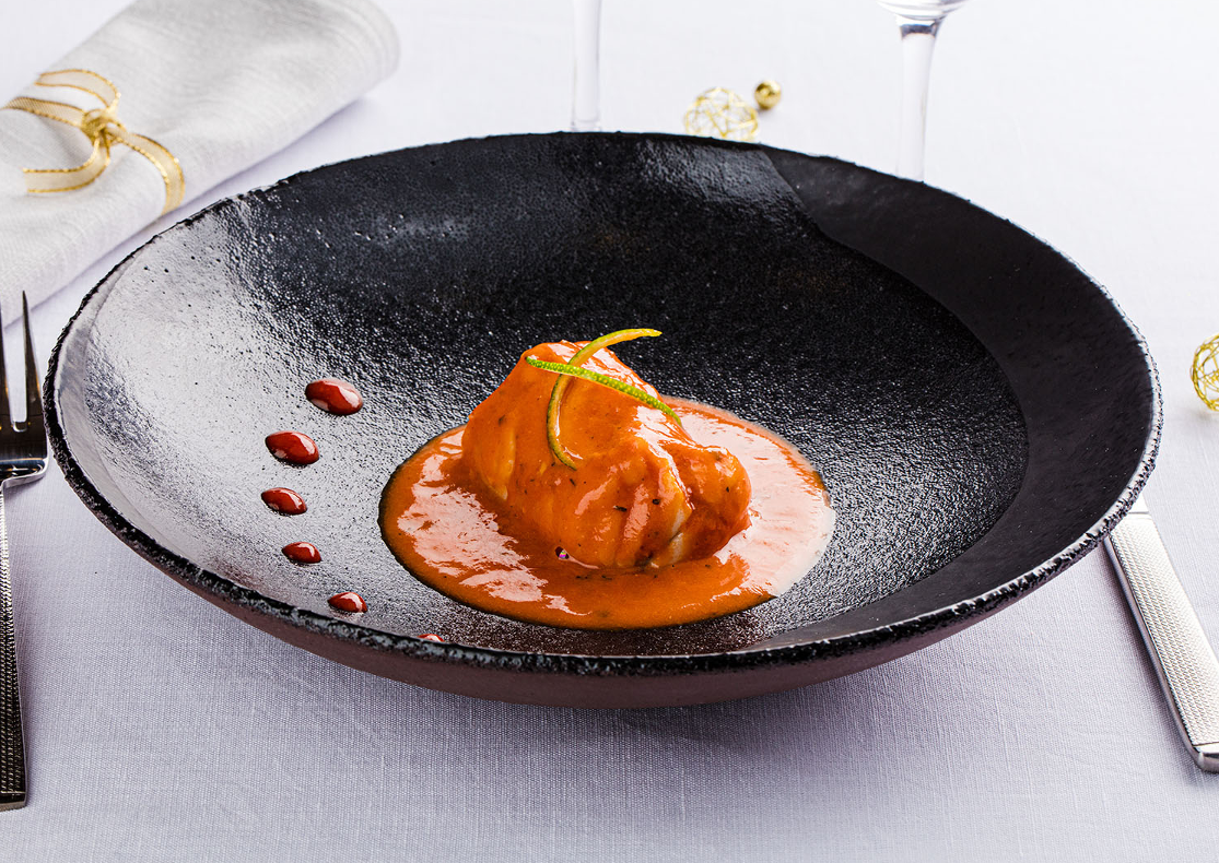 Un filet de lotte recouvert d'une sauce à l 'américaine de couleur orange, présenté dans une assiette creuse ronde et noire et agrémentée de zestes de citron vert