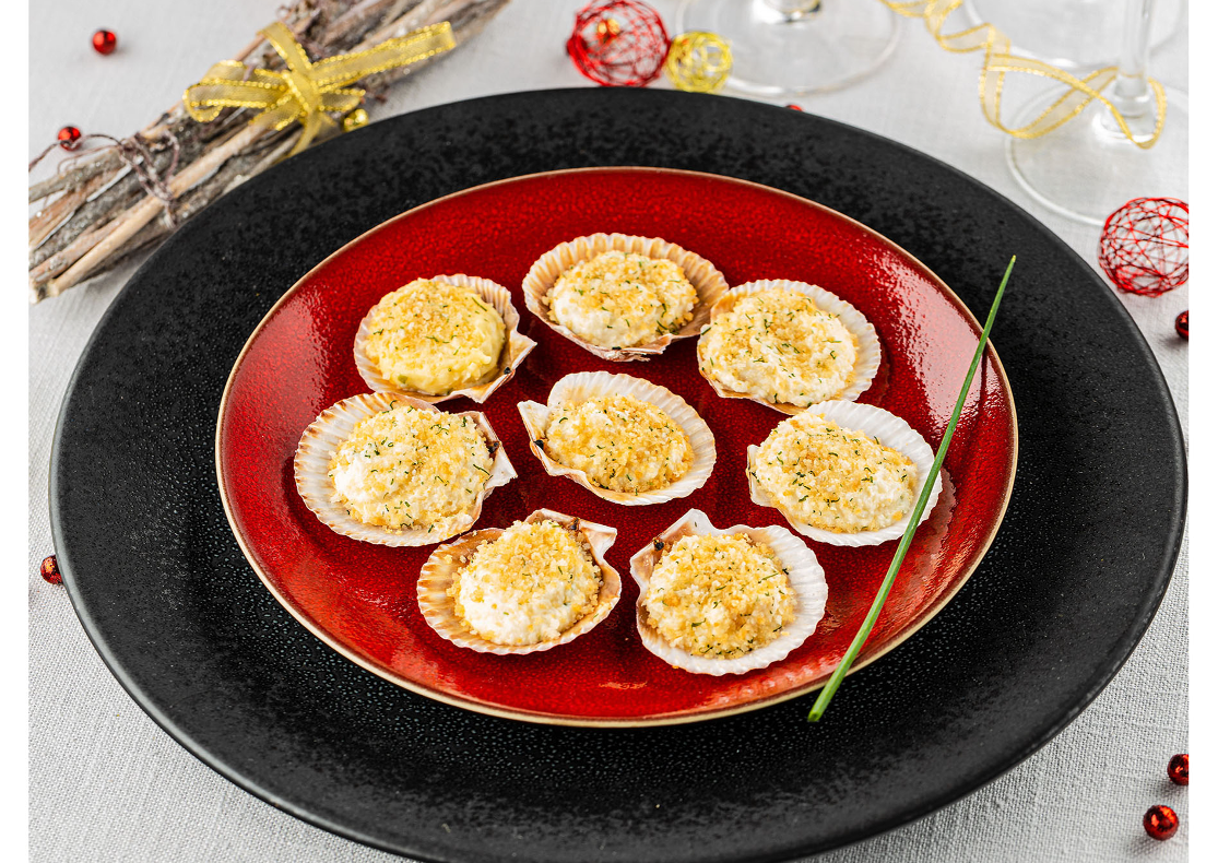 8 mini coquilles aux noix de Saint-Jacques agrémentées d'une couche de chapelure, présentées dans une assiette ronde rouge avec une branche de ciboulette posée sur le côté